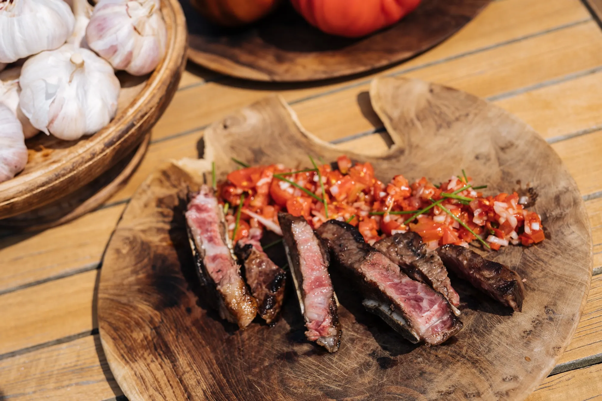 Gesneden bavette met fijngesneden tomaat op een houten plank, naast een mand knoflook en tomaten