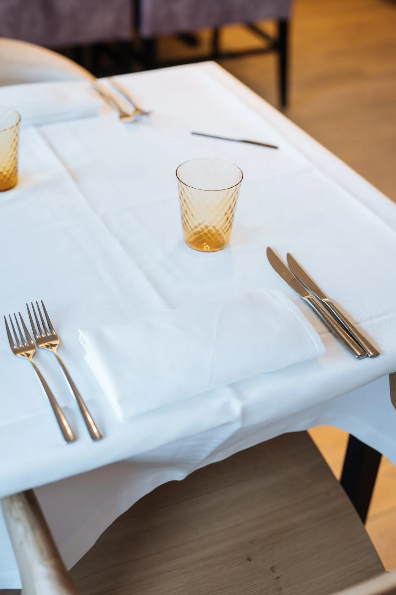 Een gedekte tafel van bovenaf: wit linnen, bestek en een amberkleurig glas