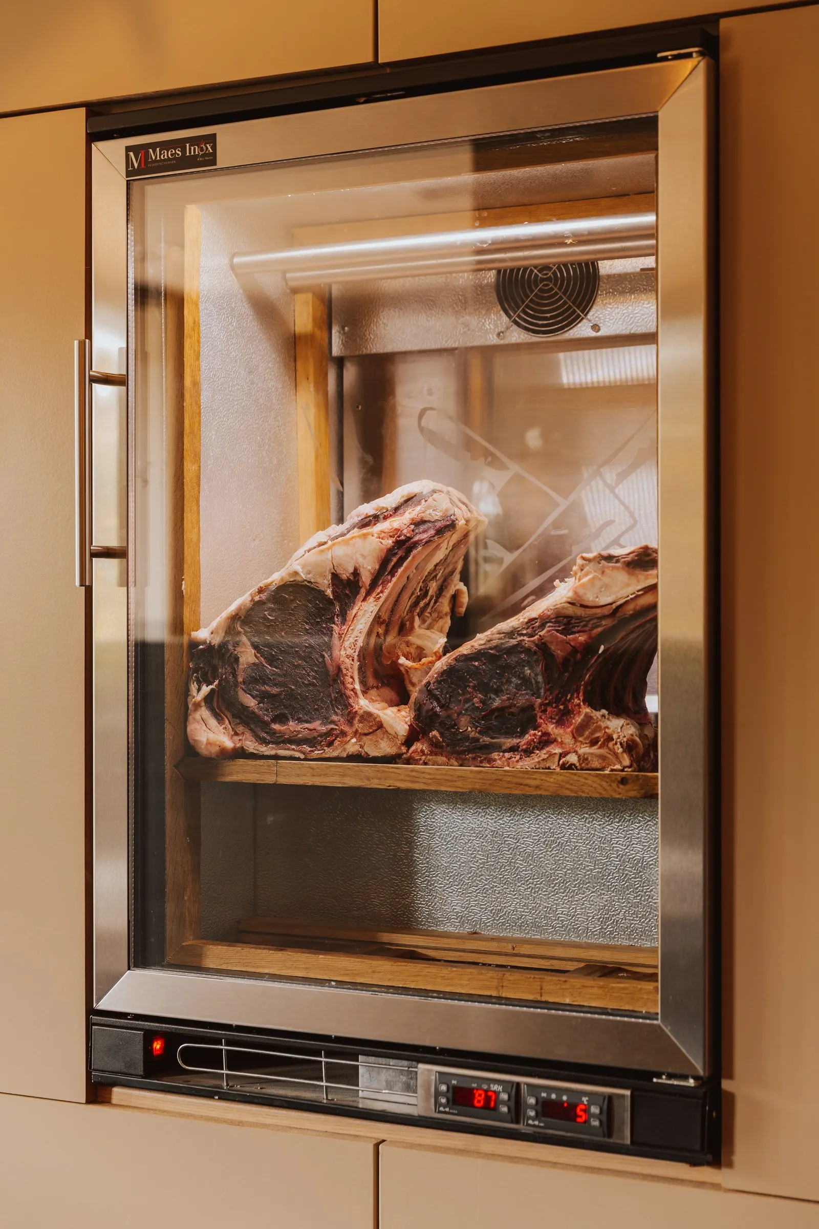 De rijpkast in de wand van het restaurant, met twee donker gerijpte ribstukken achter het glas